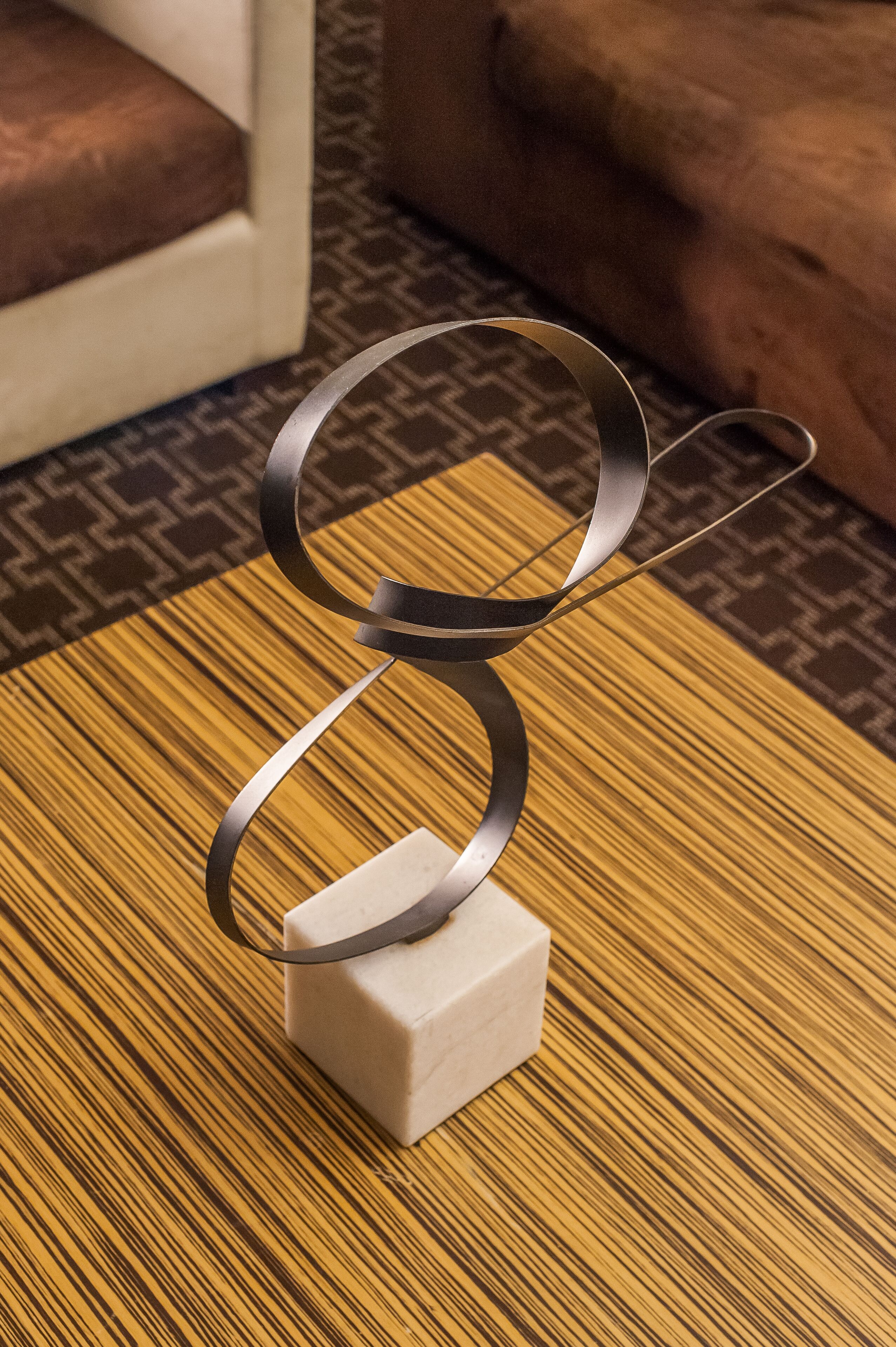 A modern metal sculpture on a wooden table with a patterned rug and a brown sofa in the background.