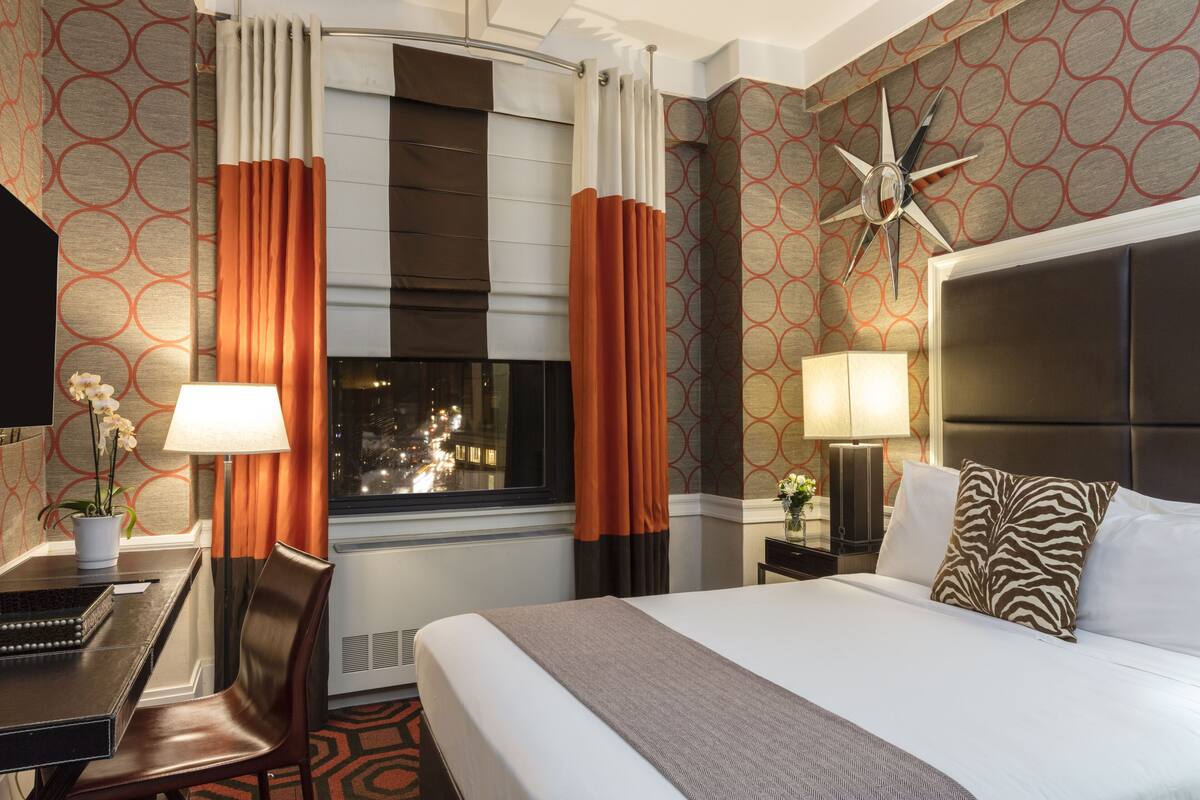 A hotel room with a bed, a desk with a chair, a lamp, a clock, and a window with curtains.