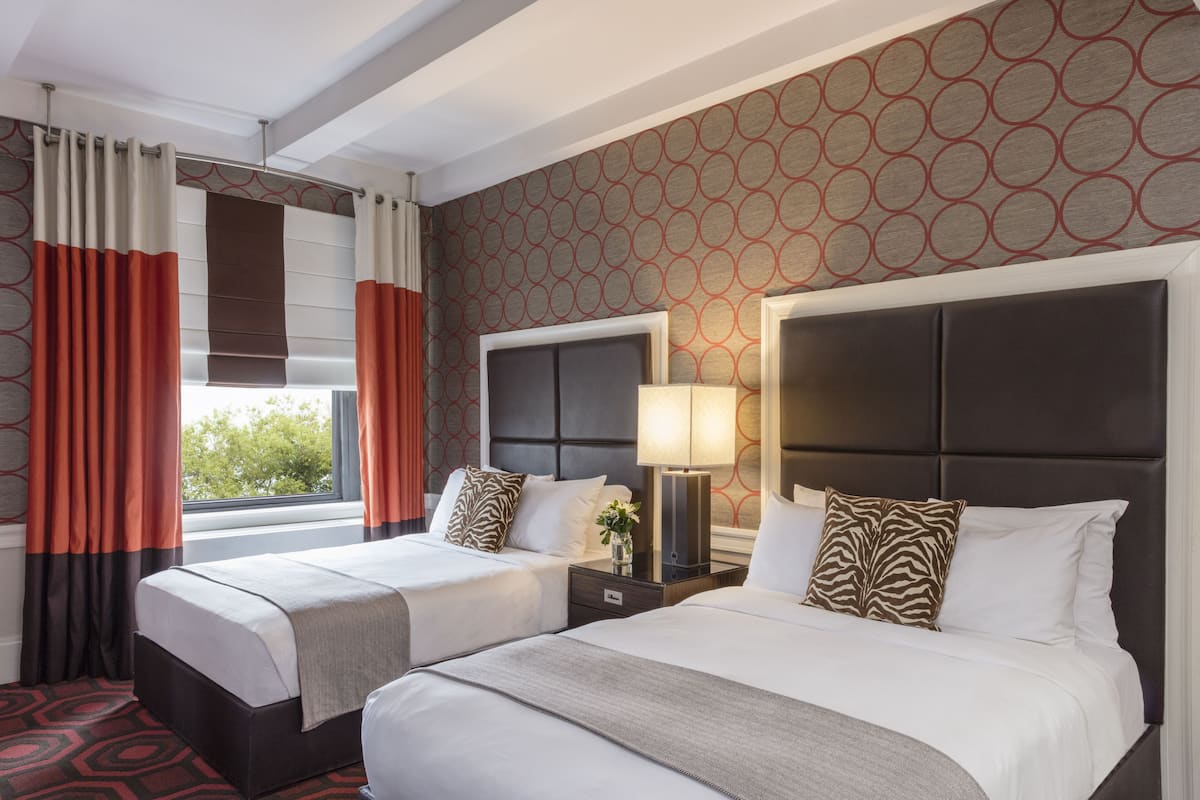 A hotel room with two beds, a large window with curtains, a headboard with a light fixture, and a patterned wall.