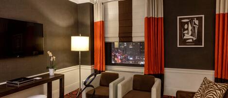Habitación de hotel con vista a la ciudad, televisor de pantalla plana, dos sillones, un sofá, una mesa de centro de madera, una mesita auxiliar y una lámpara.