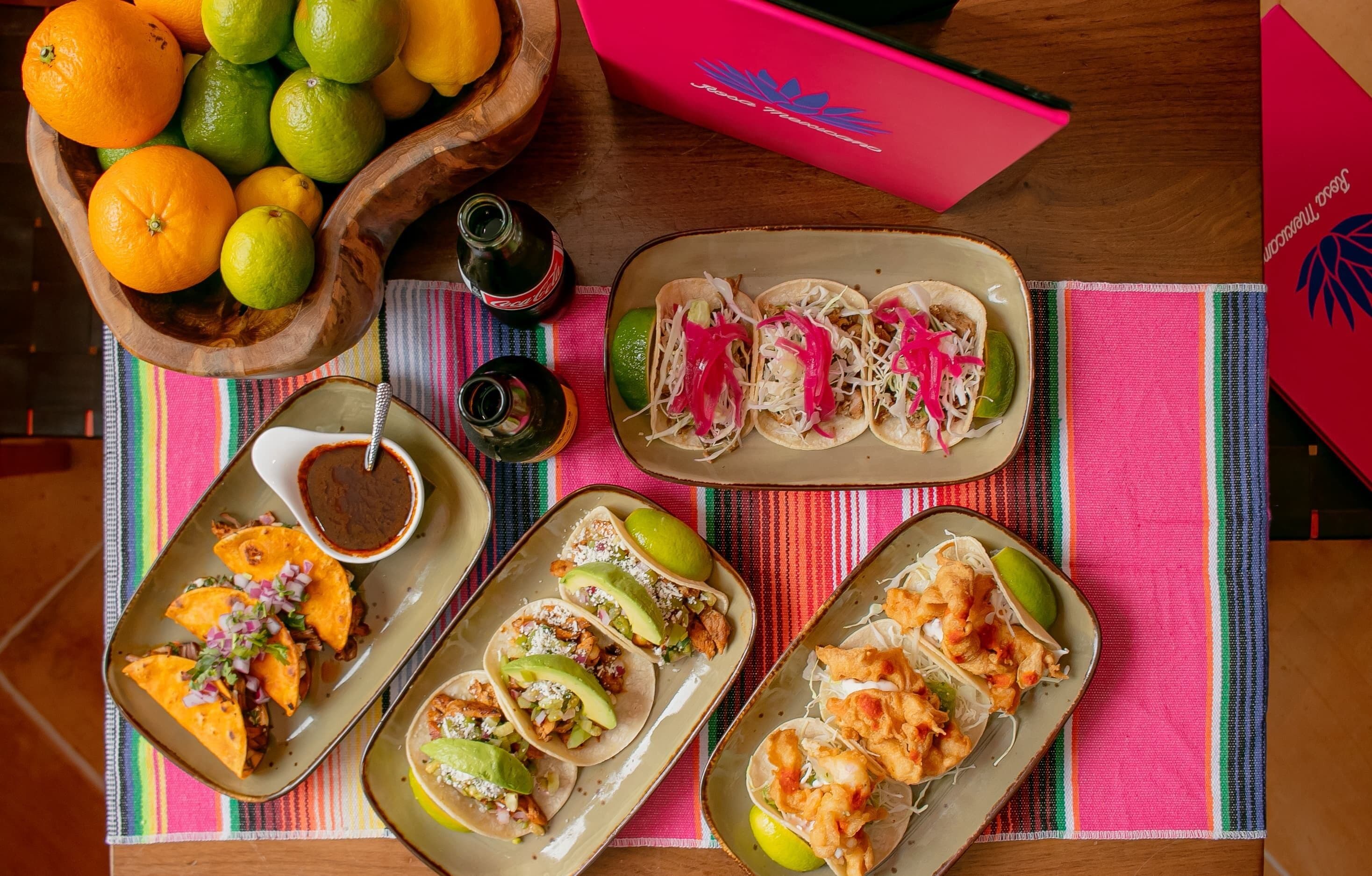 Una mesa con diferentes platillos, entre ellos tacos y un tazón de frutas.