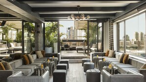 A modern lounge area with grey sofas, round ottomans, and a central coffee table. Large windows offer views of the cityscape.