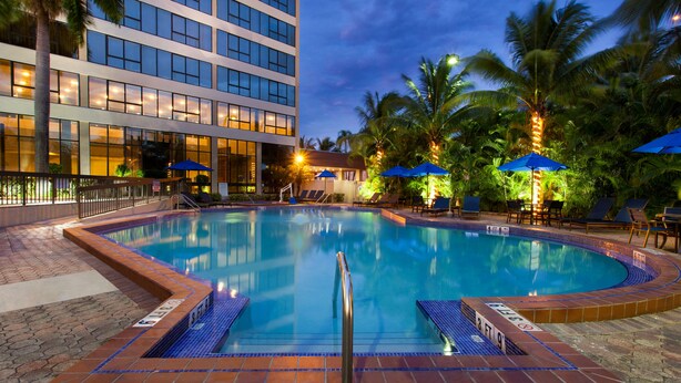 A swimming pool with a tiled edge, surrounded by lounge chairs and umbrellas, and a building with multiple floors in the background. - Holiday Inn Miami West - Airport Area by IHG (Hialeah)