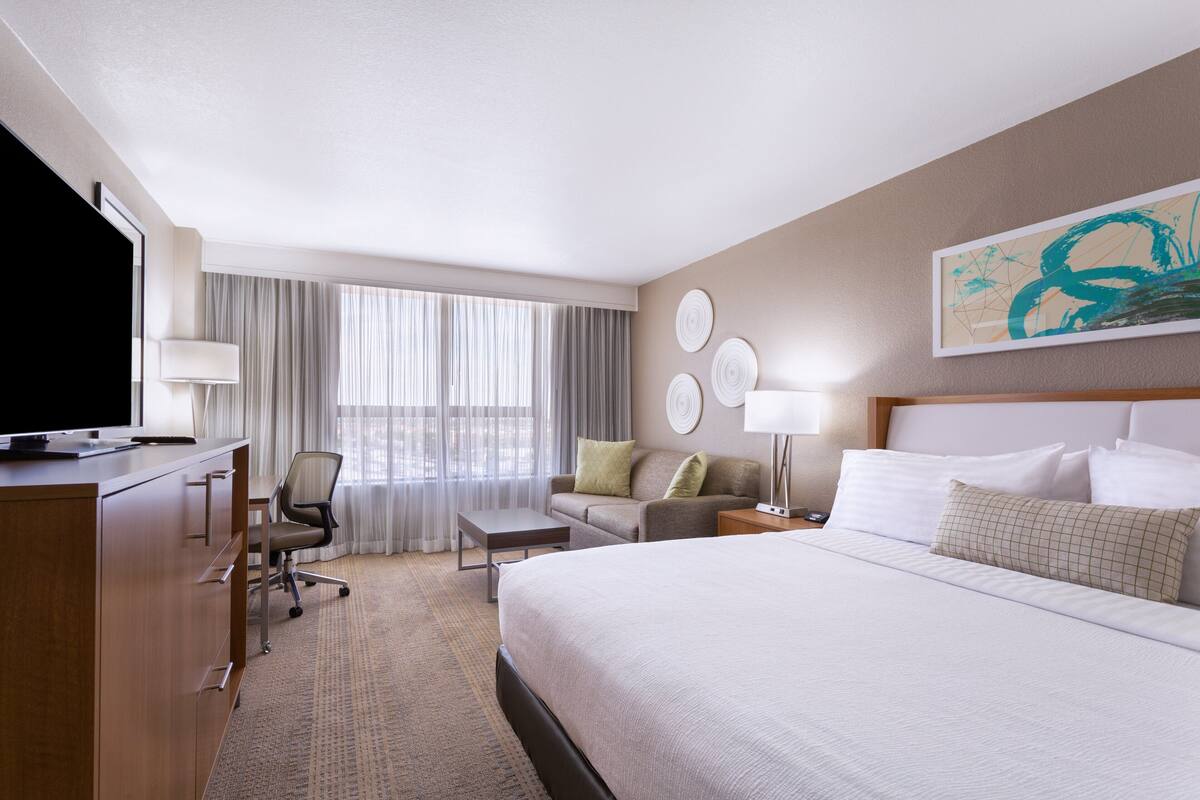 A hotel room with a bed, a desk, a chair, a TV, and a window with curtains.