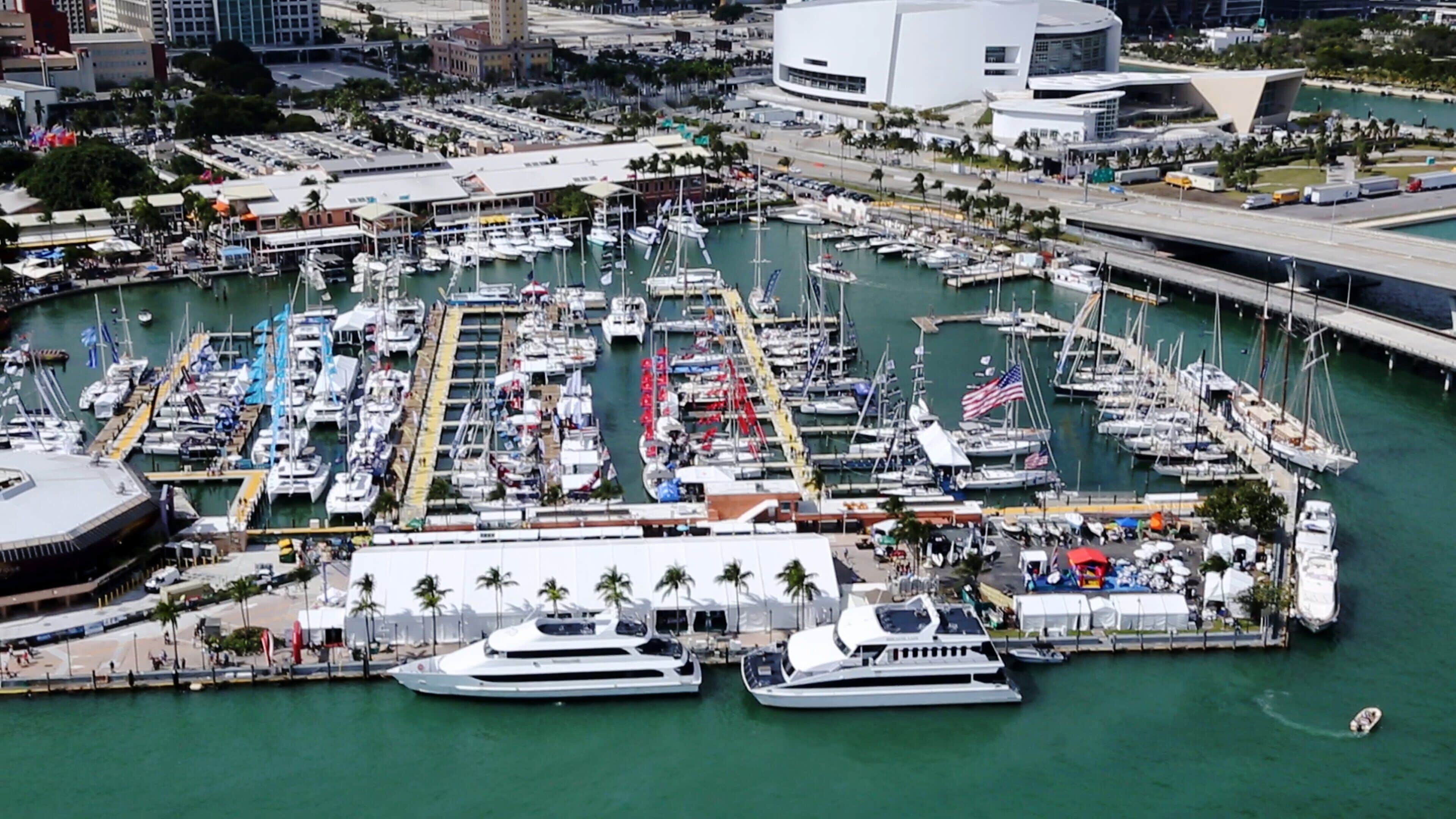 A marina with multiple boats docked, a large white building, and a bridge in the background.
