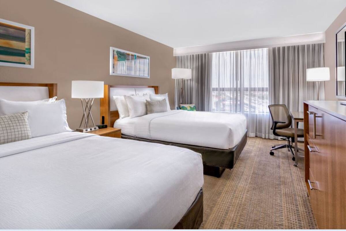 A hotel room with two beds, a desk, a chair, and a large window with curtains.