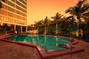 Uma piscina com um deck ladrilhado, cadeiras de descanso e palmeiras.