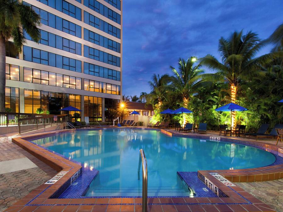 A swimming pool with lounge chairs and umbrellas, surrounded by palm trees and a hotel building.