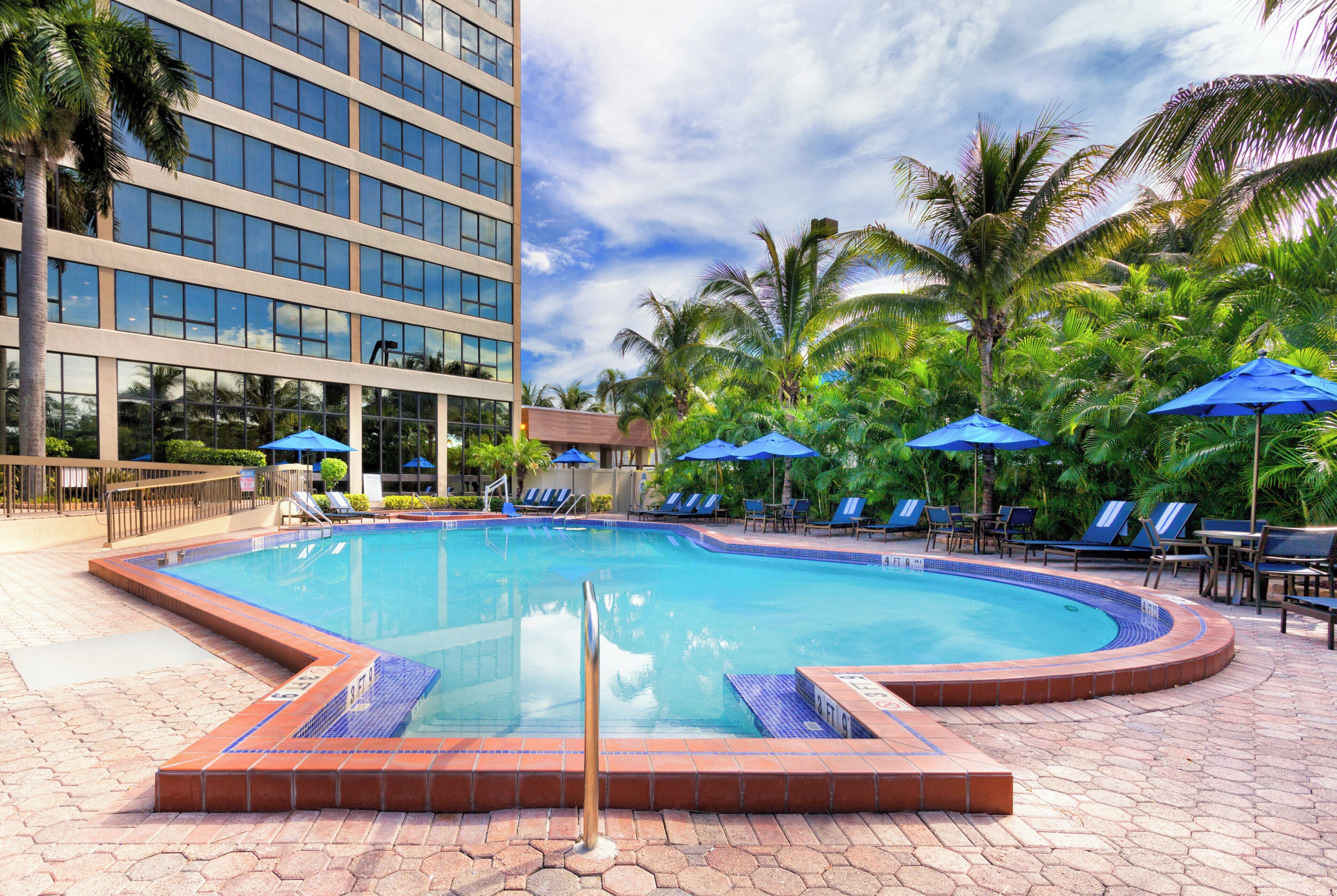Una piscina con borde de azulejos, rodeada de sillones de descanso y sombrillas, con un edificio al fondo.