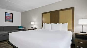 A hotel room with a large bed, a gray sofa, a small table with a lamp, and a wall-mounted painting.