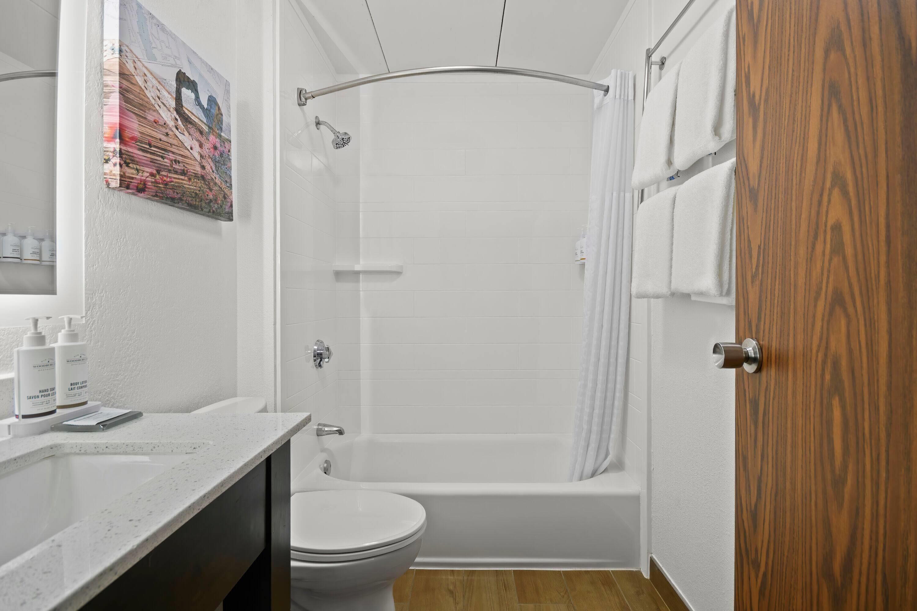 Een badkamer met een wit bad, toilet en een douchegordijn. Er is een houten deur en een wit wastafelmeubel met een spiegel.