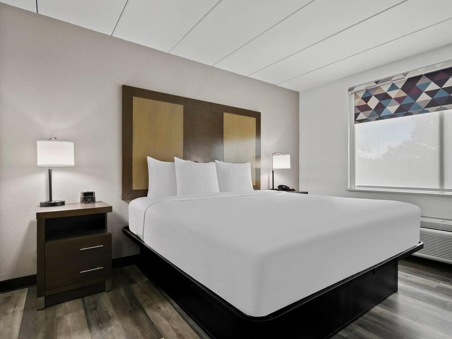Habitación de hotel con una cama grande, una mesita de noche con lámpara, un calentador en la pared y una ventana con cortina de diseño geométrico.