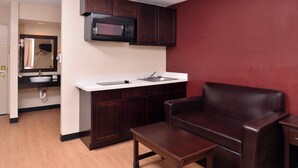 Una kitchenette compacta con microondas, lavamanos y gabinete, una pequeña mesa de madera y un sillón de cuero.