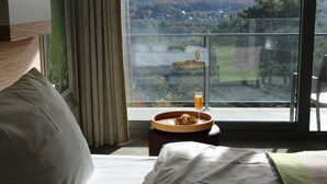 Habitación de hotel con cama, mesita auxiliar con bandeja de pasteles y vaso de jugo de naranja, y un ventanal que ofrece vistas a un jardín y colinas.