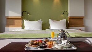 Una bandeja de desayuno bien dispuesta con croissants, pasteles y bebidas, sobre una cama con cabecera verde y almohadas blancas.