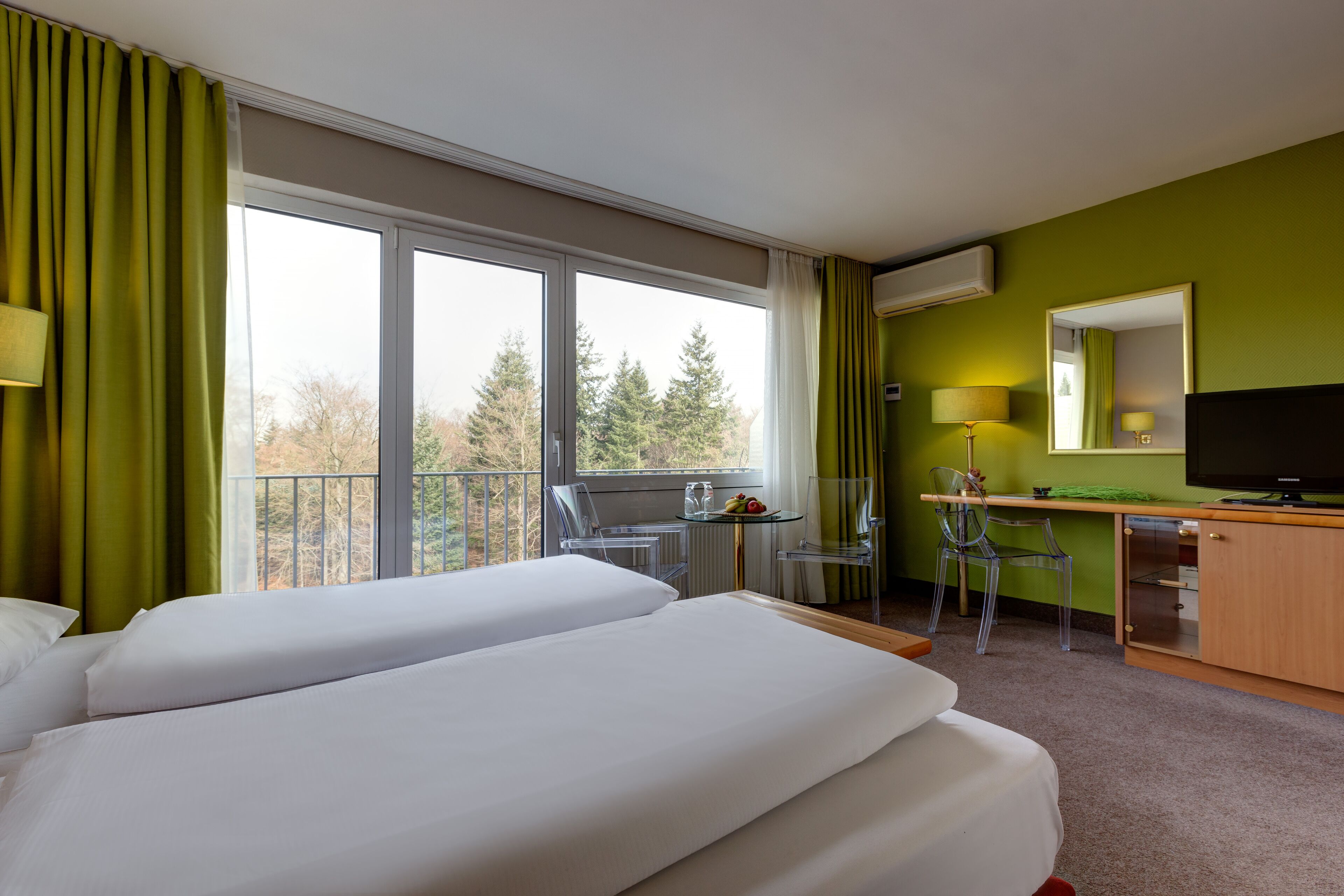 A hotel room with two beds, a desk with a chair, a television, and a balcony with a view of trees.