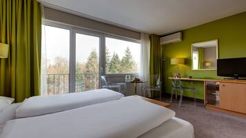 Een hotelkamer met twee bedden, een bureau met stoel, een televisie en een balkon met uitzicht op bomen.