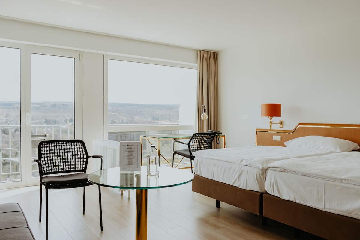 A hotel room with a bed, two chairs, a small table, a lamp, and large windows offering a city view.