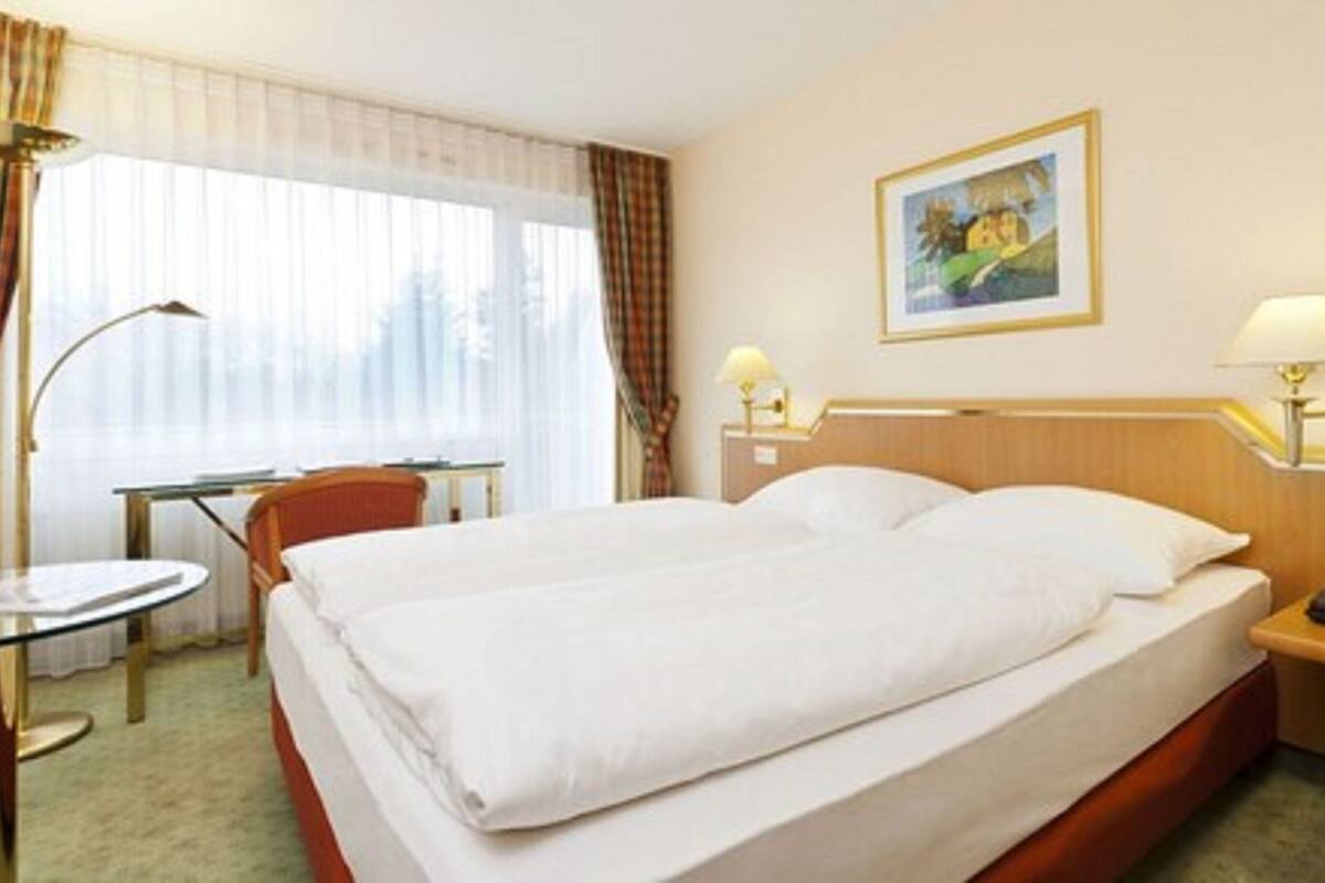 A hotel room with a large bed, a desk, a chair, a lamp, a painting, and a telephone on the nightstand.