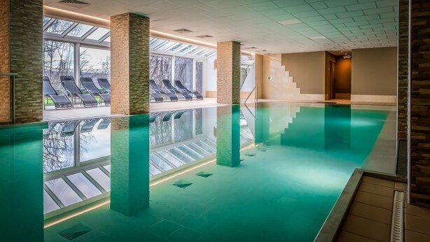Una piscina interior con paredes de vidrio transparente, suelo de baldosas y una escalera que sube a un nivel superior.