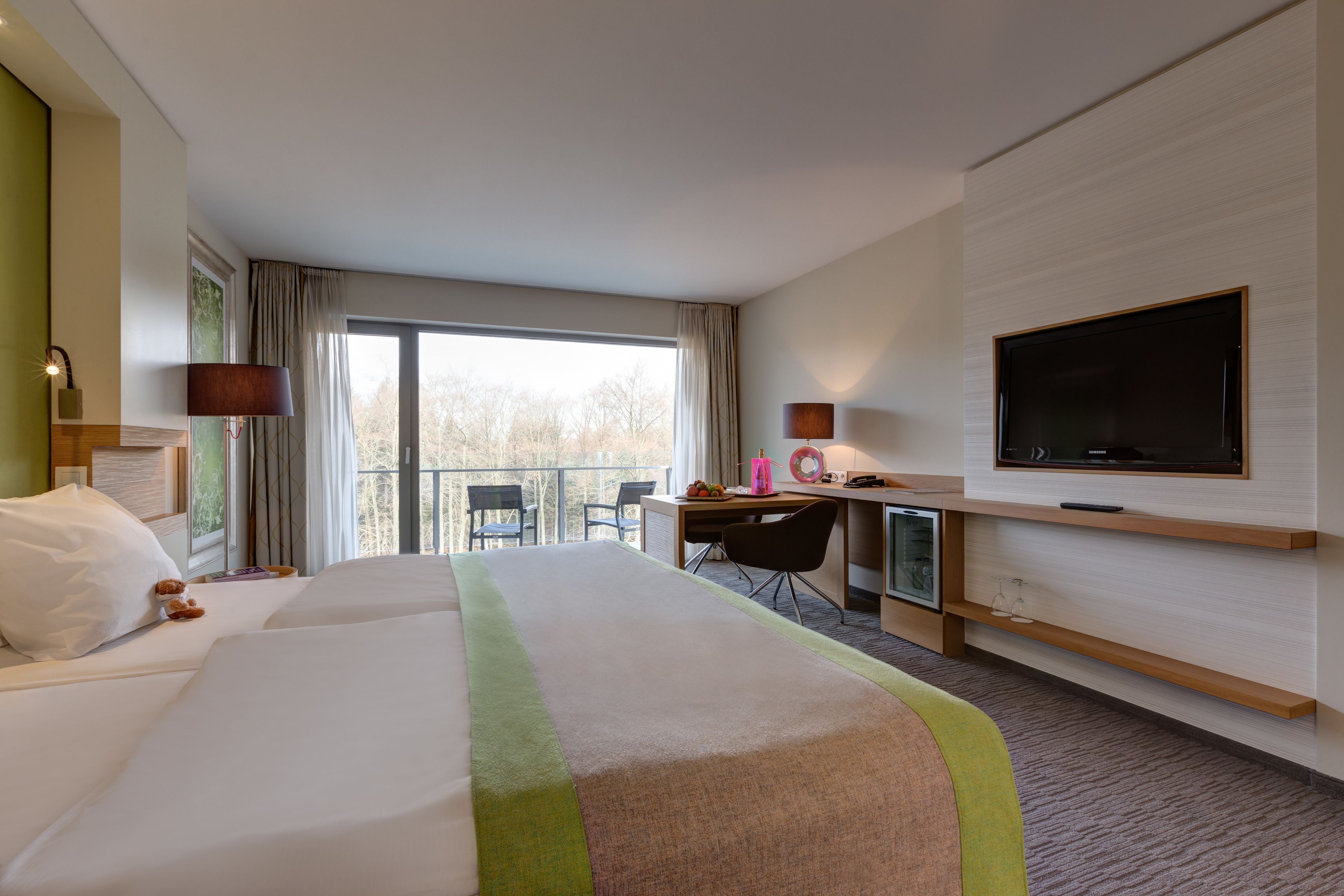 Habitación de hotel con dos camas, un escritorio, una silla, un televisor y un balcón con vistas a árboles.