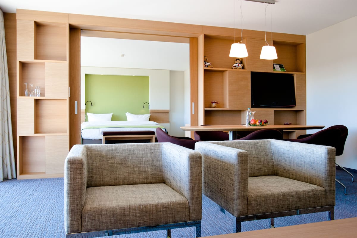 A hotel room with a bed, two armchairs, a desk with a flat-screen TV, and a wooden shelving unit with various items.