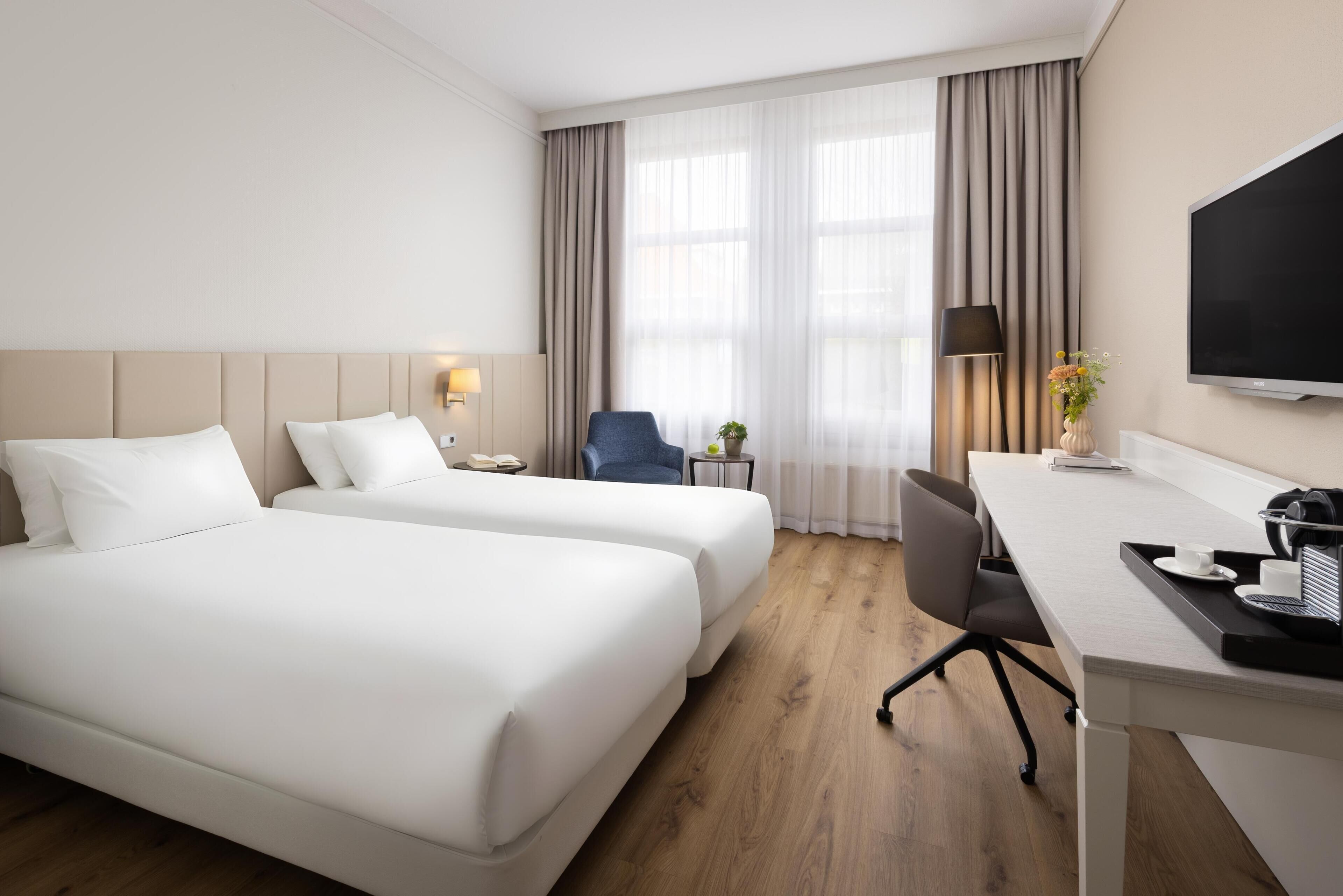 A hotel room with two beds, a desk, a chair, a TV, and a window with curtains.