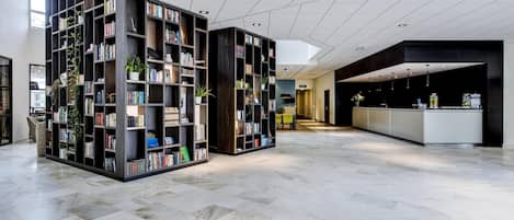A spacious lobby with a modern design, featuring a large central area with a reception desk, surrounded by tall bookshelves filled with books and plants.