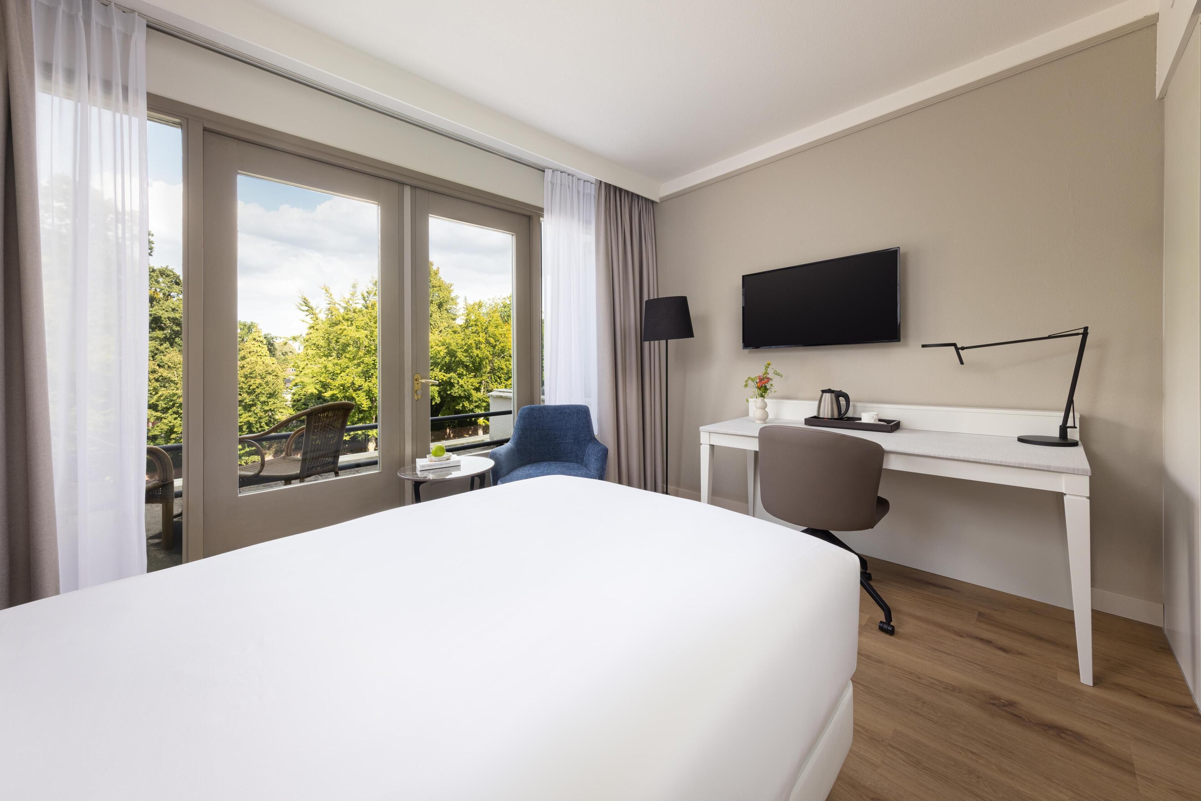 Une chambre d’hôtel avec un lit, un bureau, une chaise, une télévision et un balcon donnant sur la verdure.