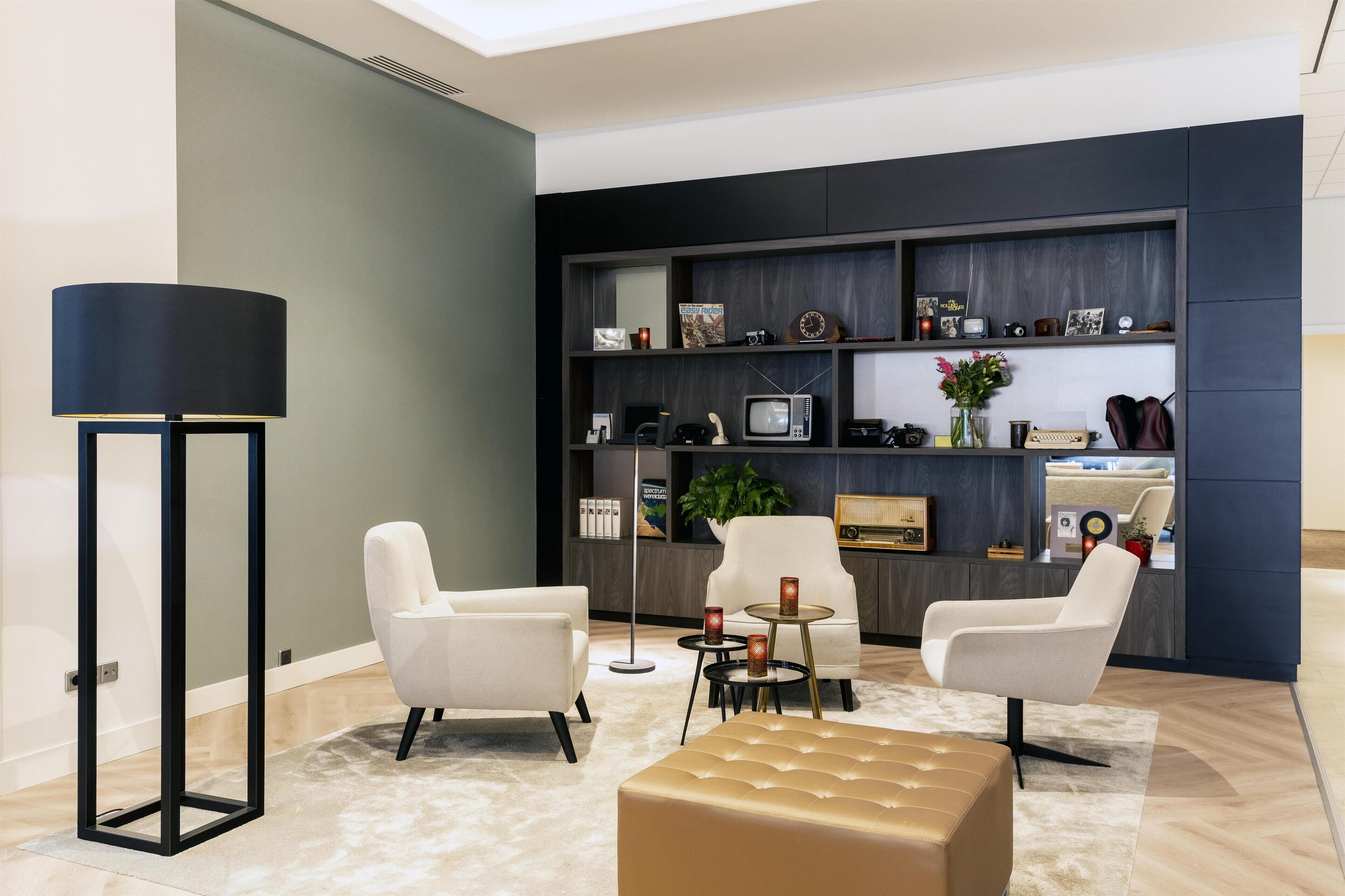 A modern living room with a sofa, armchairs, a coffee table, and a floor lamp. There is a bookshelf with various items and a small table with a plant.