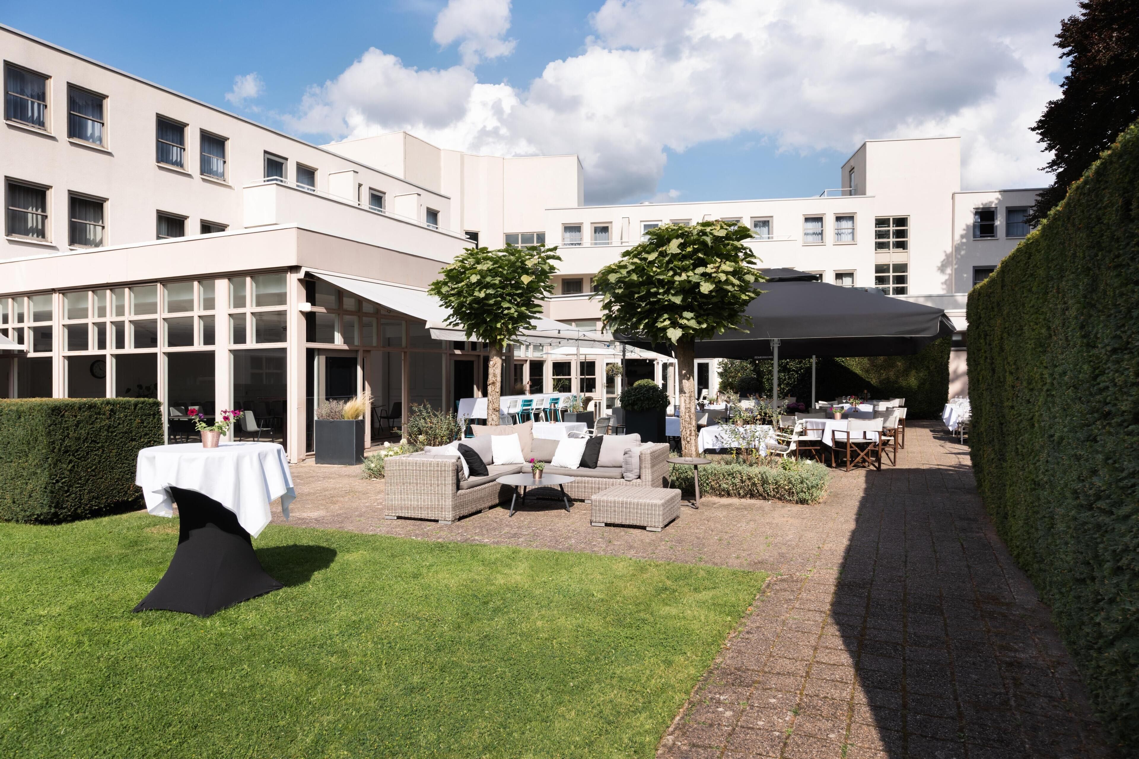 An outdoor seating area with tables and chairs, a covered patio, and a hedge.