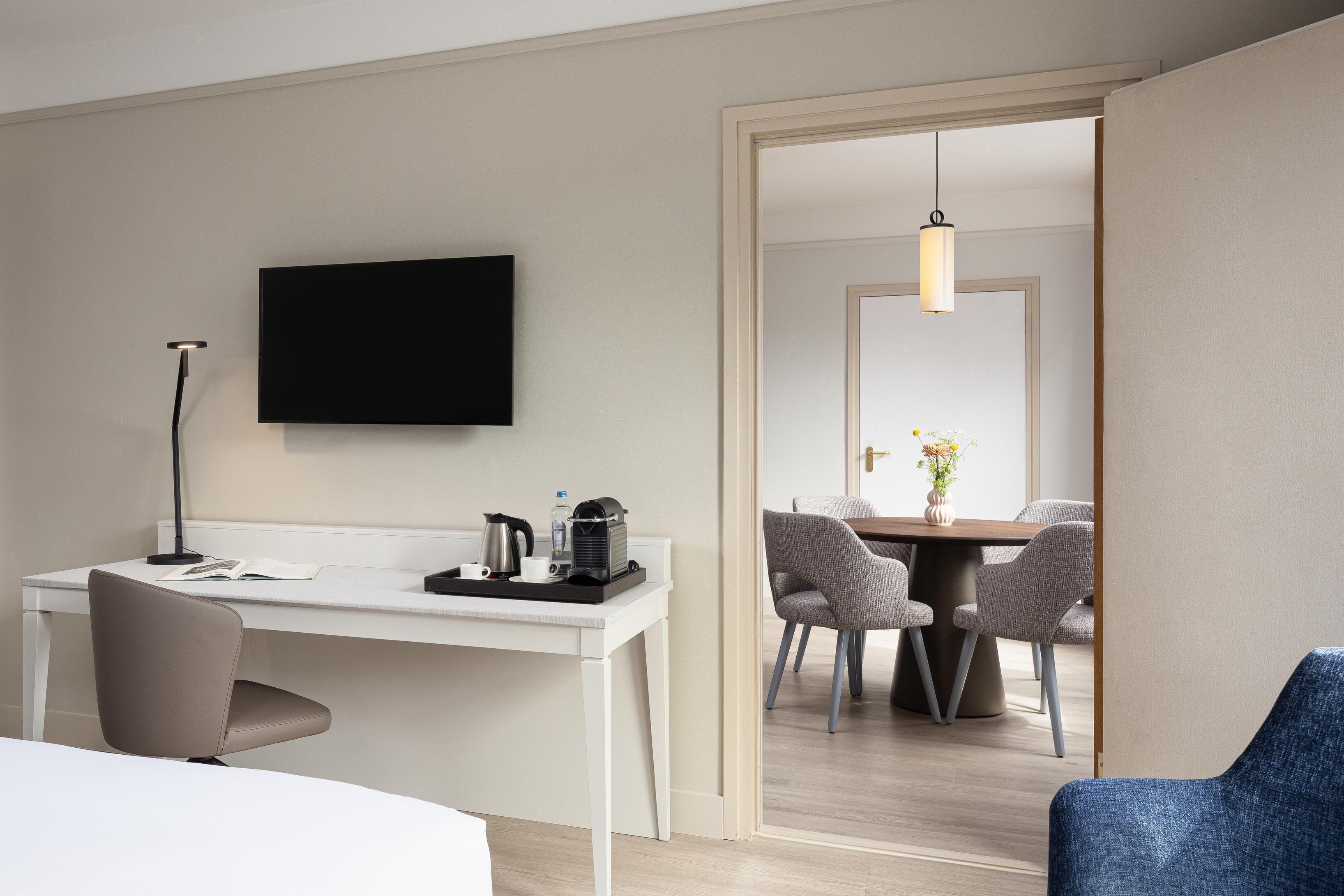 Habitación de hotel con escritorio, silla, televisor de pantalla plana, mesita de noche y zona de comedor con sillas y mesa.