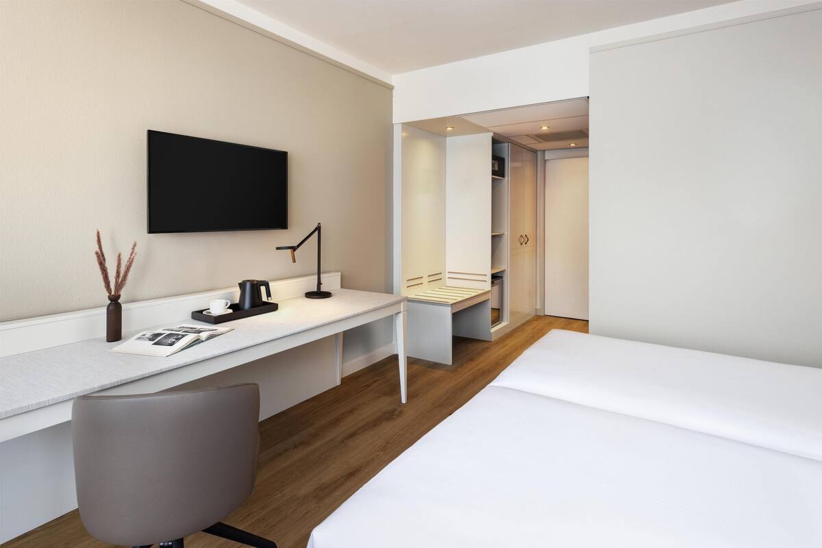 A modern hotel room with a white desk, a chair, a bed, and a wardrobe.