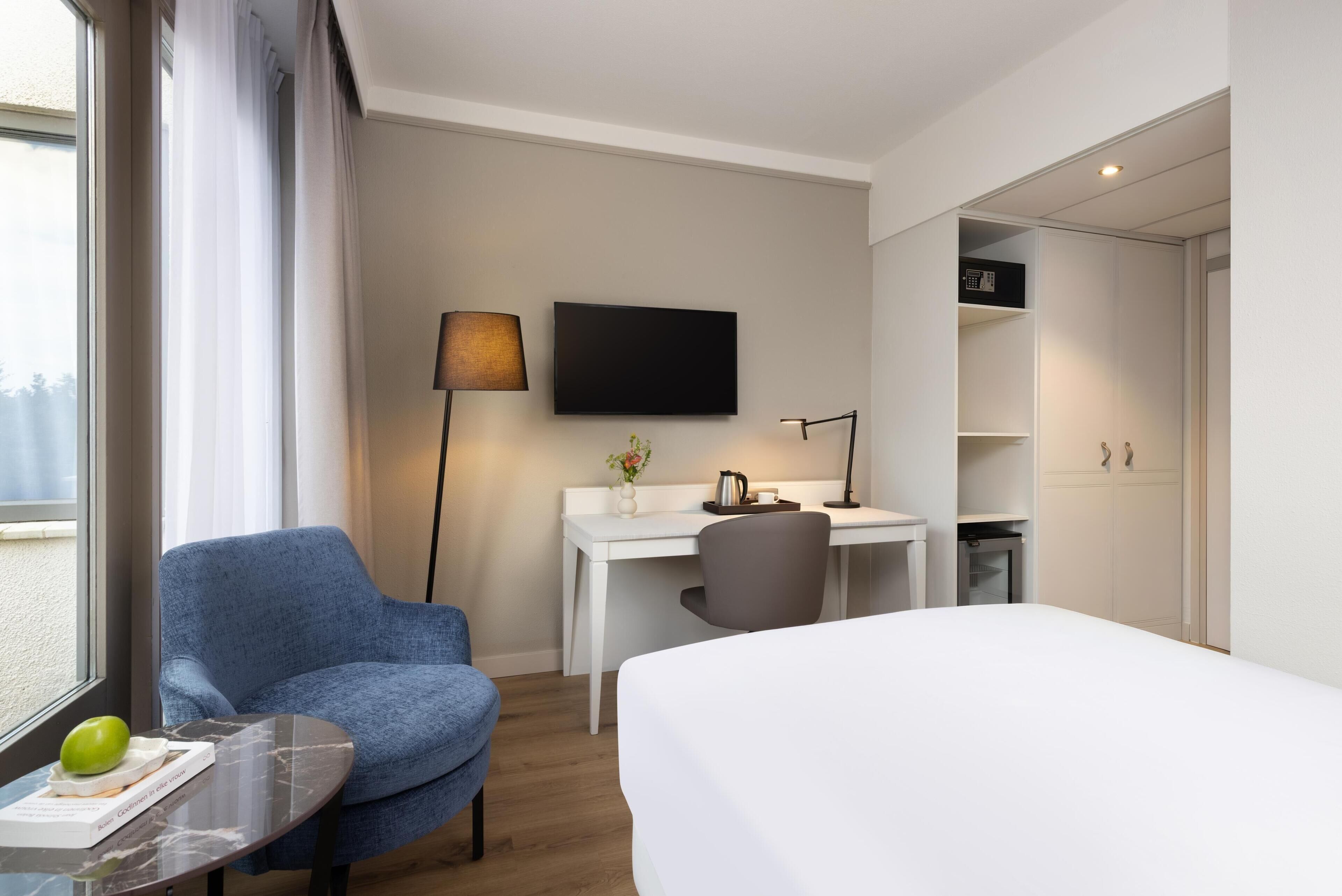 Una camera d'albergo con un letto, una scrivania, una sedia, una TV e una finestra con tende.