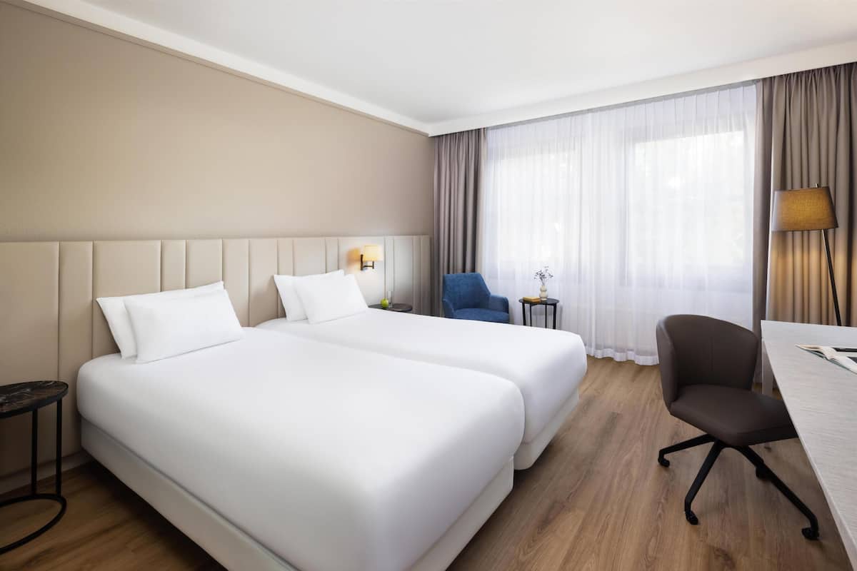 A hotel room with two beds, a desk, a chair, a lamp, and a window with curtains.
