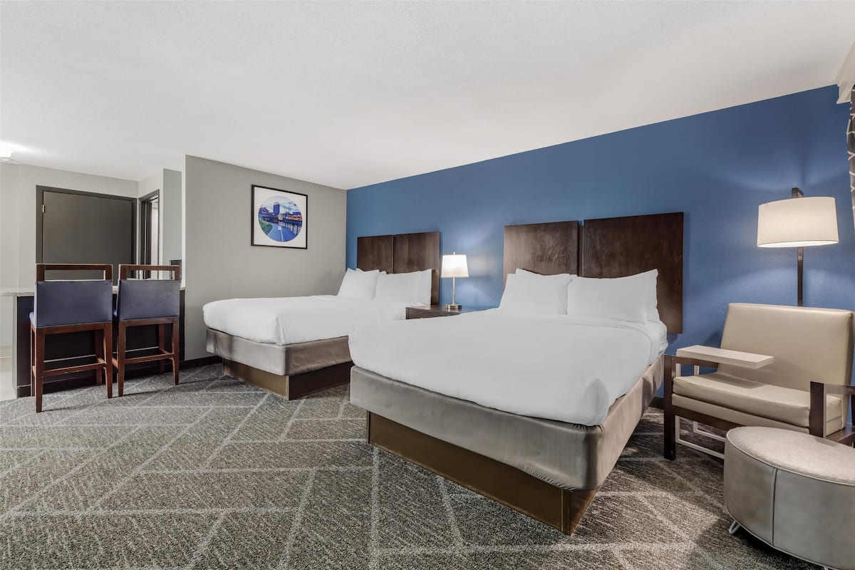 A hotel room with two beds, a desk, chairs, and a lamp.