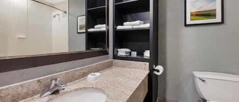 A bathroom with a granite countertop sink, a mirror, a toilet, and a shelf with folded towels.