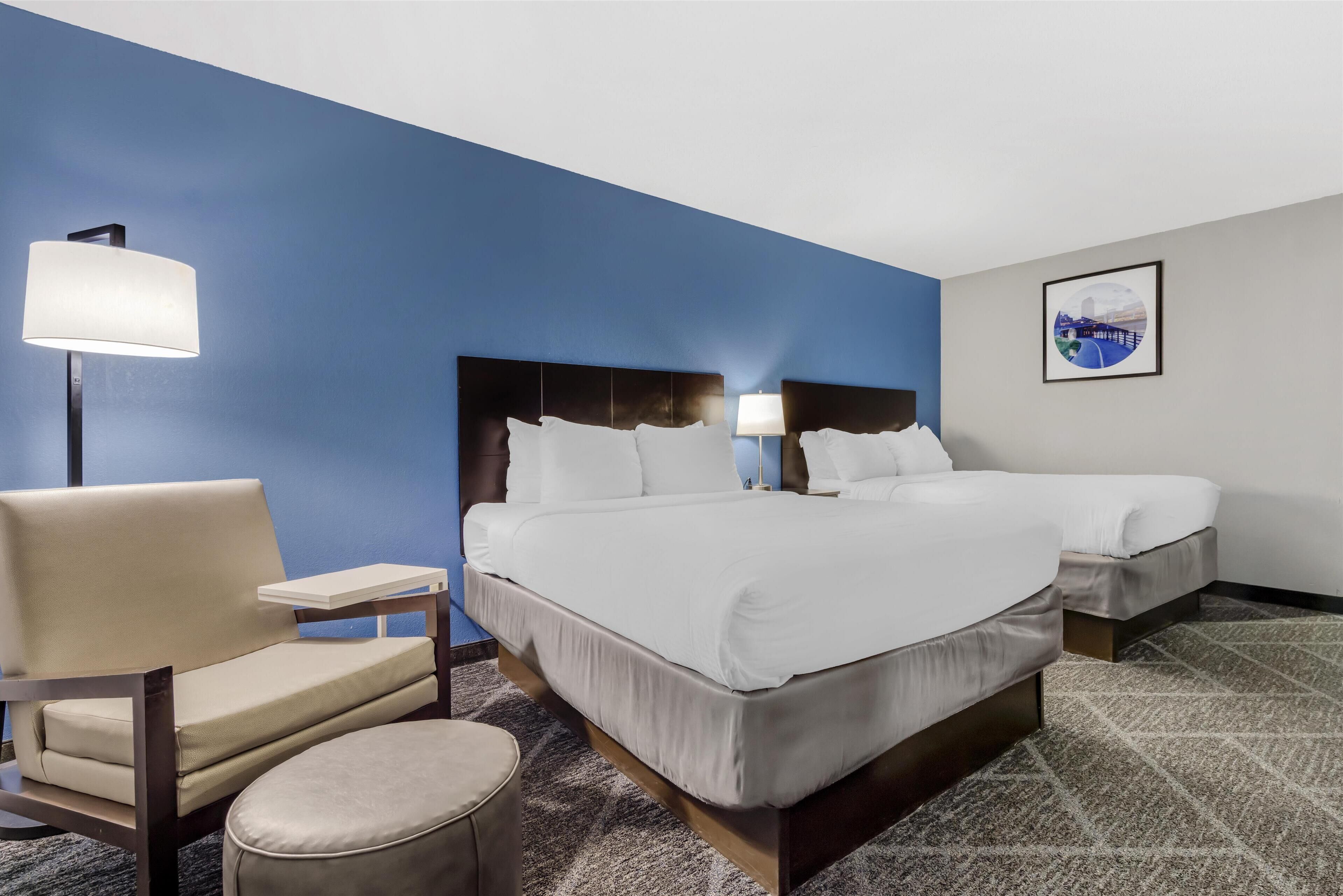 A hotel room with two beds, a chair, a lamp, and a framed picture on the wall.