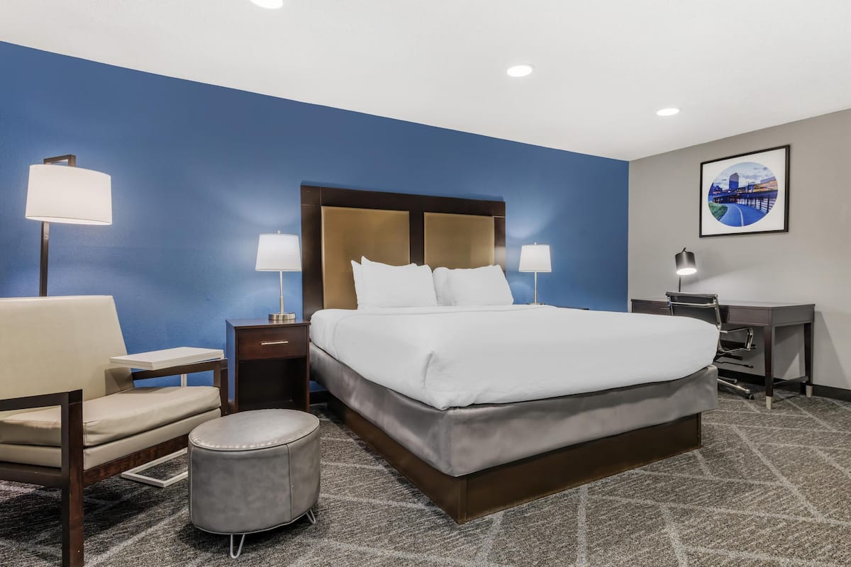A hotel room with a large bed, two bedside tables with lamps, a desk, a chair, and a round ottoman.