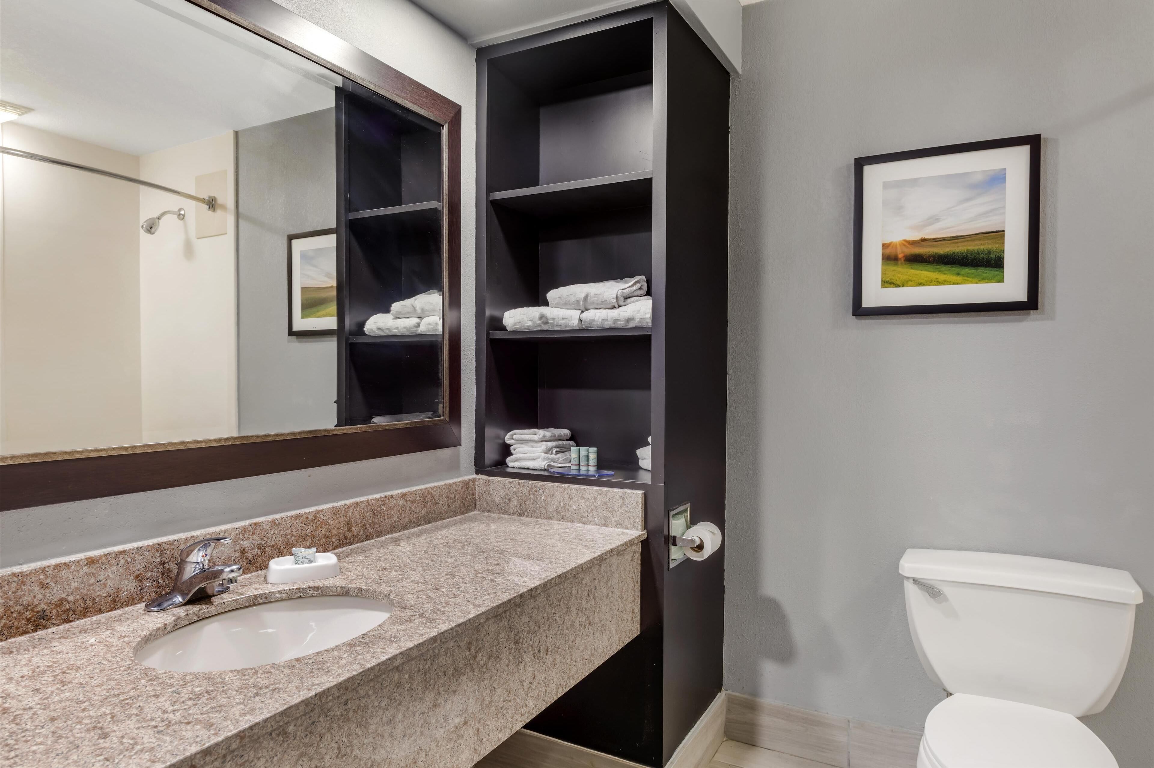 Une salle de bain avec un lavabo doté d’un plan de toilette en granit, un grand miroir, des toilettes et une étagère avec des serviettes pliées.