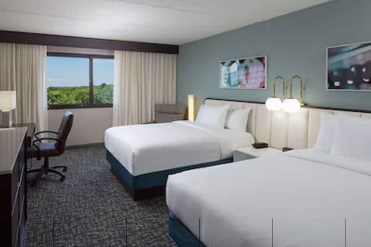 A hotel room with two beds, a desk with a flat-screen TV, a chair, and a window with curtains.