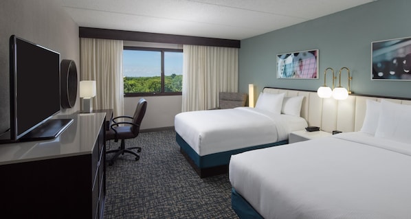 Quarto de hotel com duas camas, uma escrivaninha com computador, uma cadeira, janela com cortinas e decoração na parede.