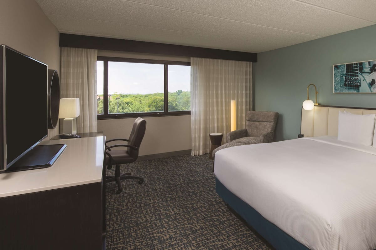 A hotel room with a large bed, a desk with a laptop, a chair, a desk lamp, a bedside table, and a window with curtains.