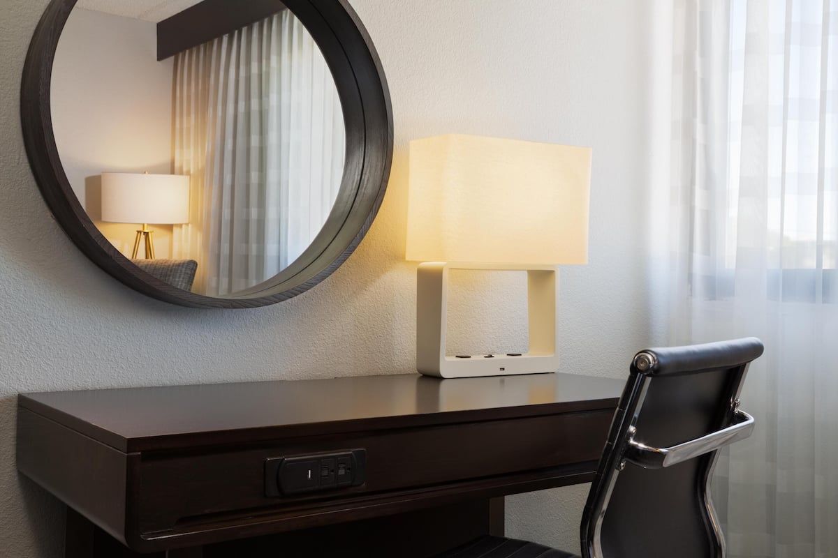 A hotel room with a desk, a lamp, a chair, and a round mirror.