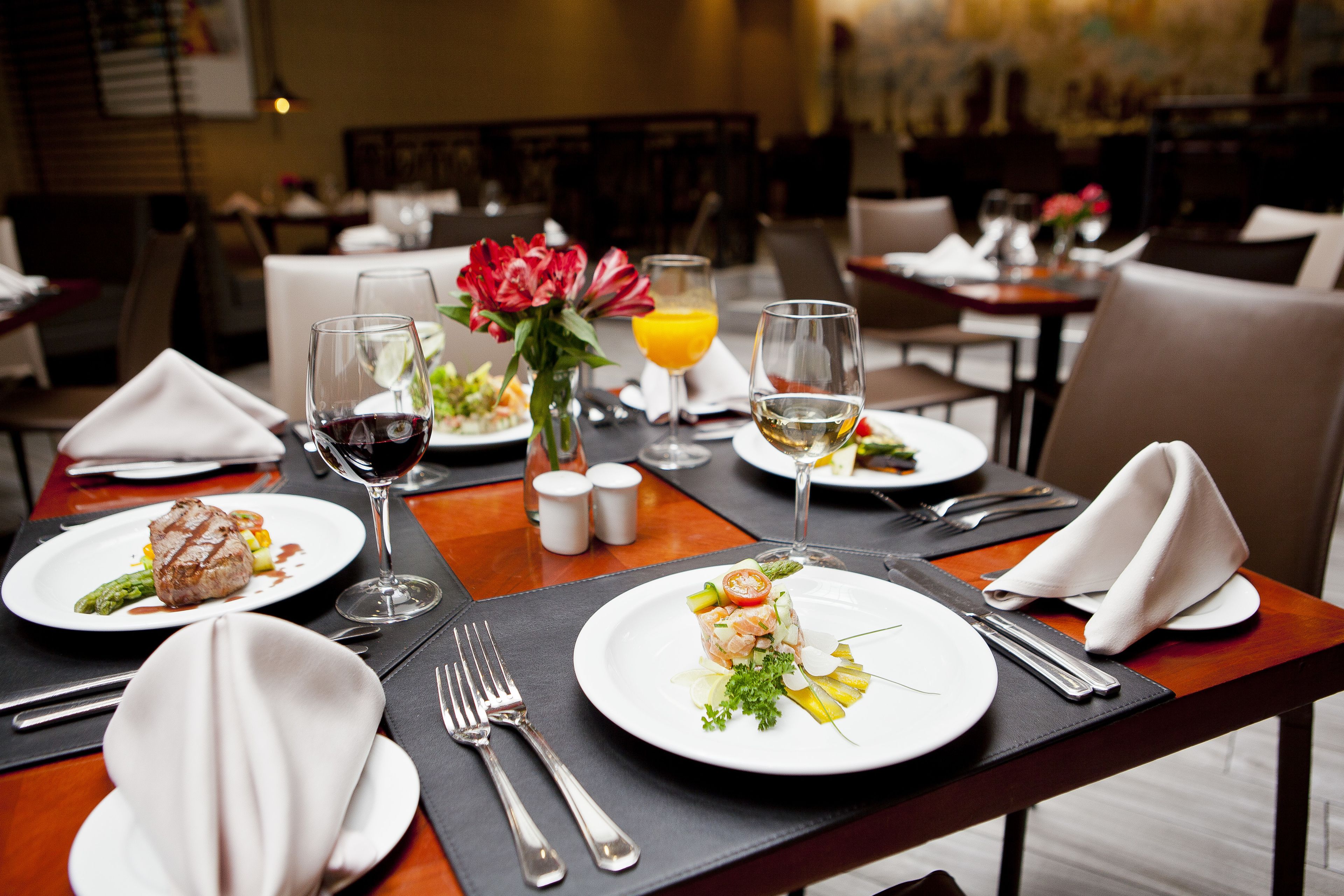 Una mesa de comedor dispuesta con platos de comida, copas de vino y un florero con flores.