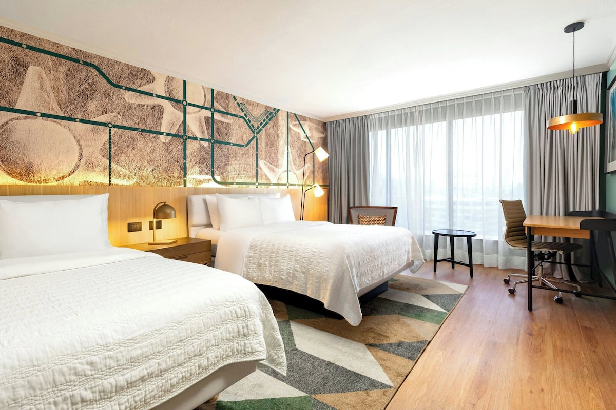 Habitación de hotel con dos camas, un escritorio, una silla, una lámpara y un ventanal grande con cortinas.