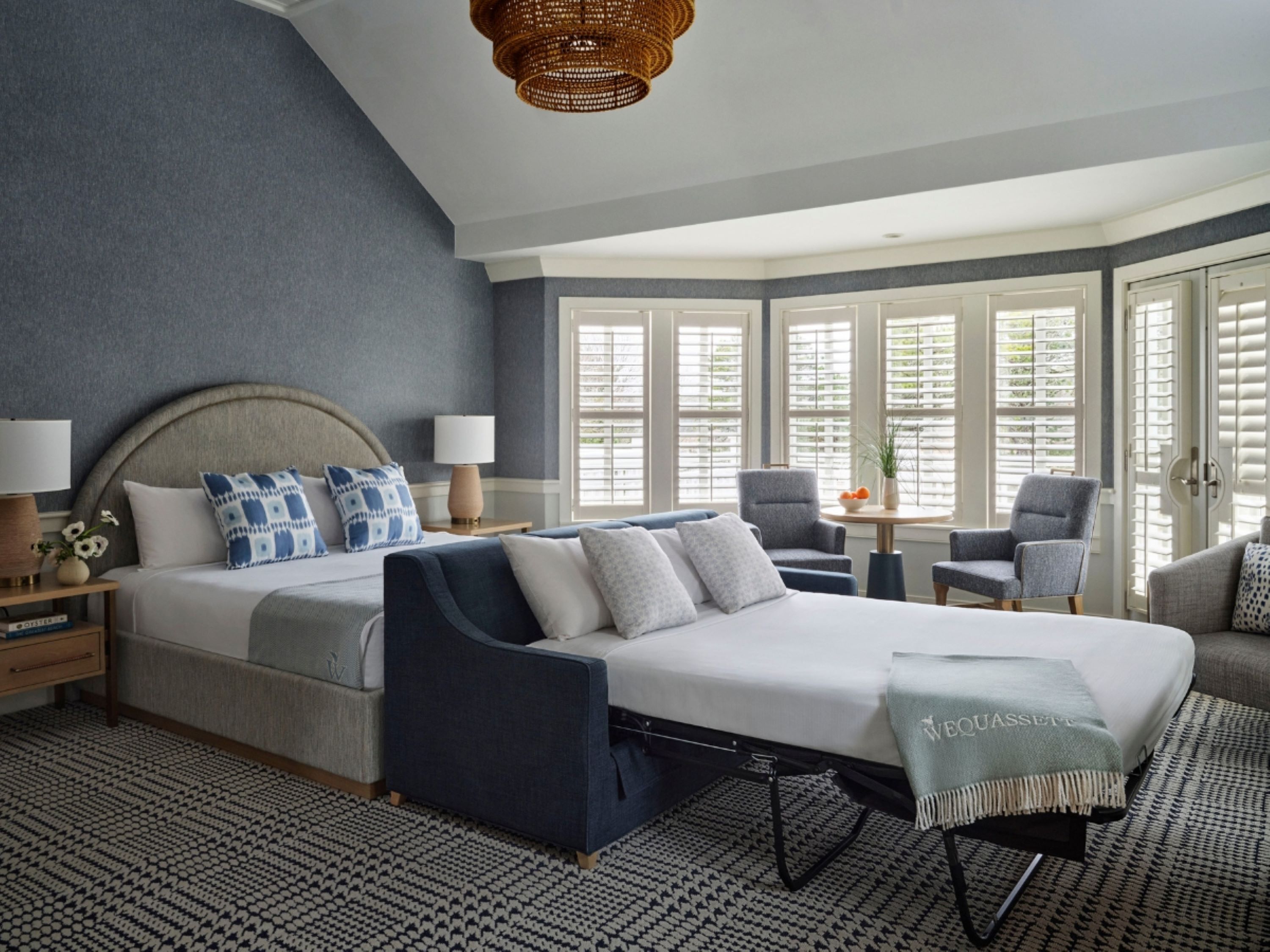 Une chambre à coucher avec un grand lit, deux tables de chevet, une chaise, une petite table et une fenêtre avec des stores.