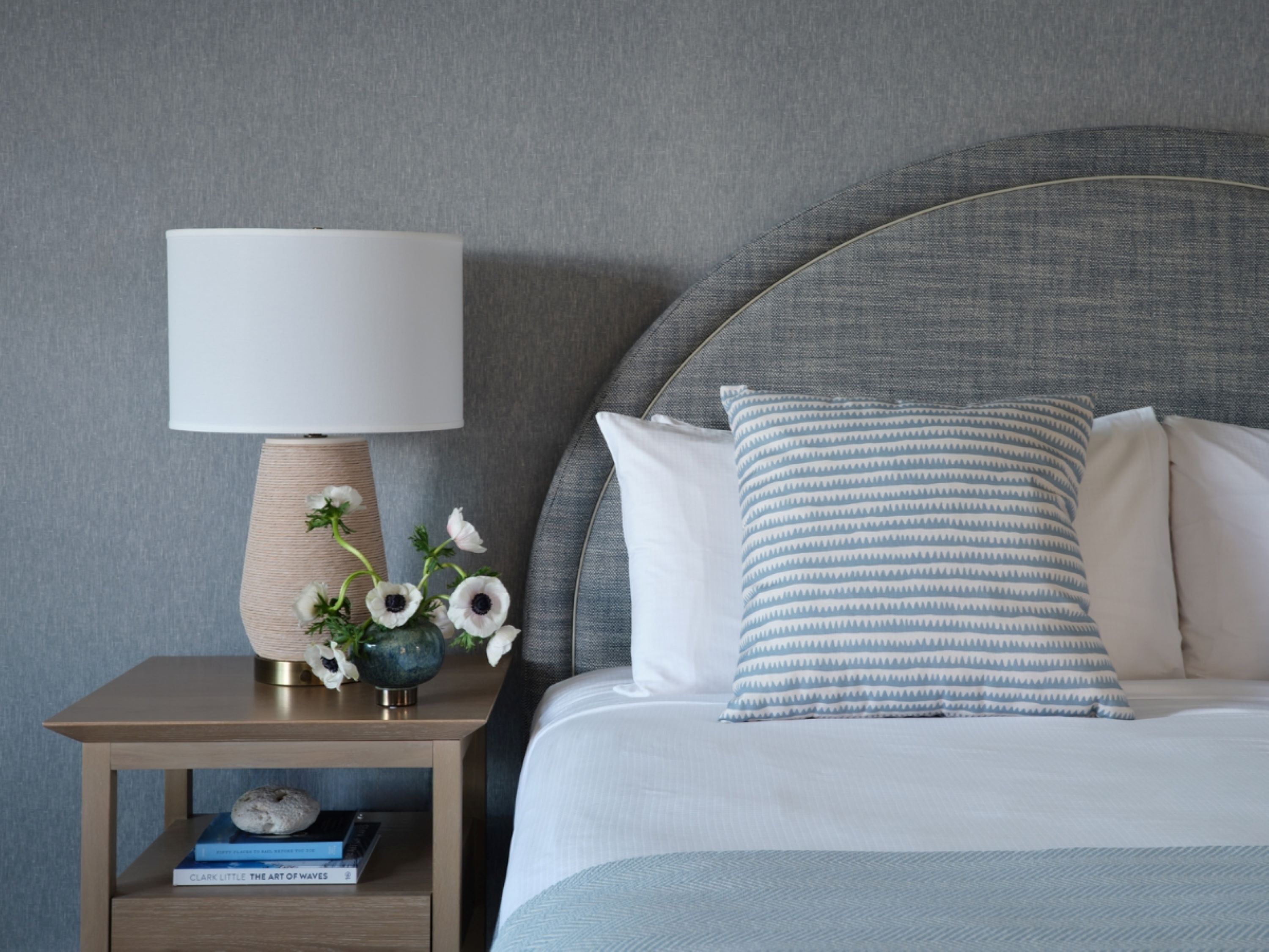 A neatly arranged bedroom with a bed, pillows, a bedside table with a lamp, and a small shelf with books.