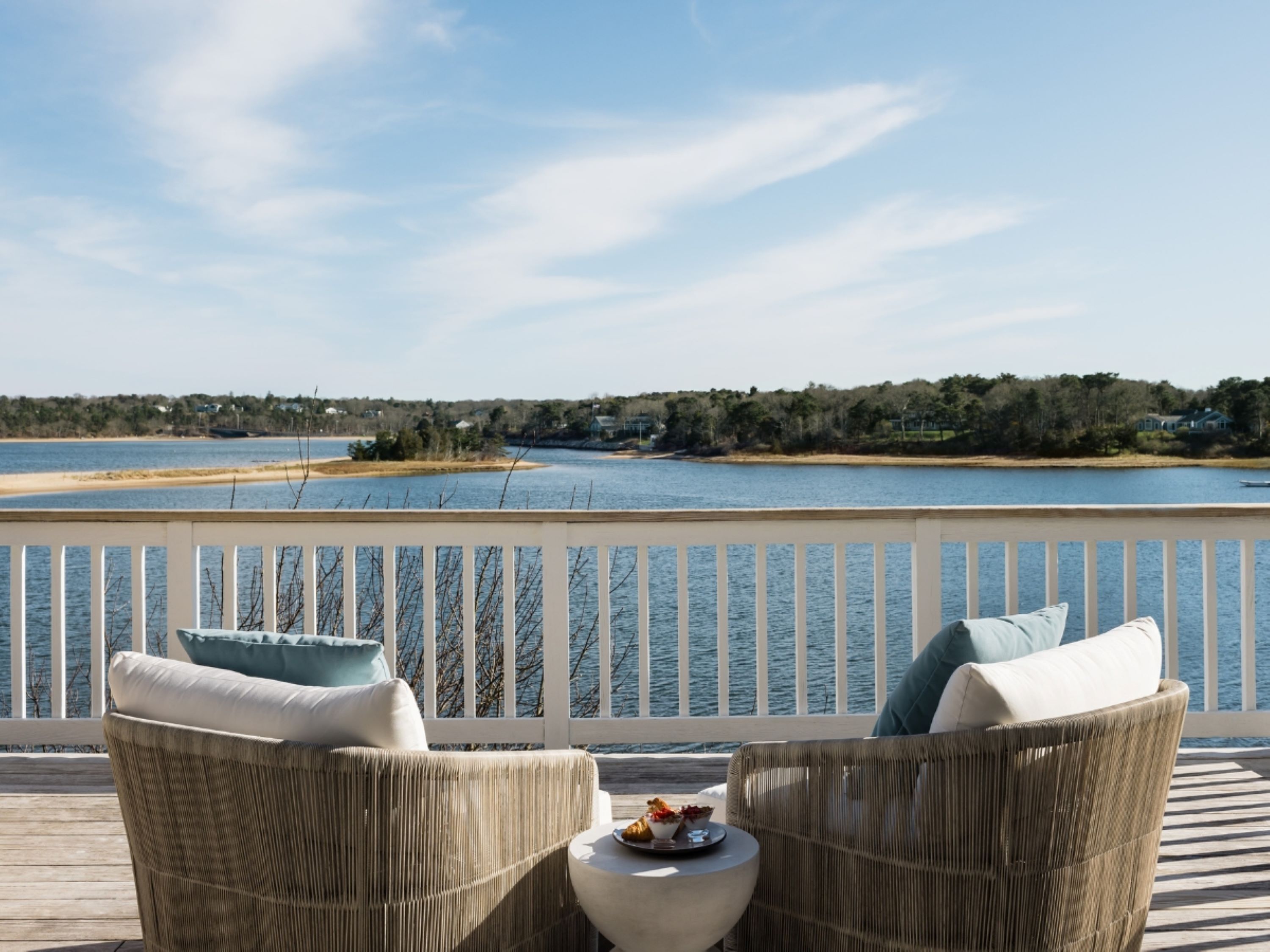 A deck with wicker chairs and a small table, overlooking a lake with a distant shoreline.