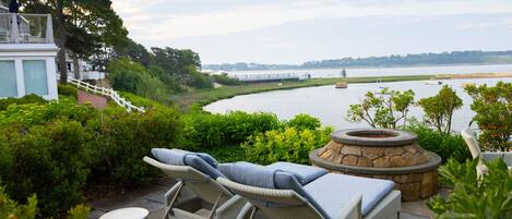 A patio with lounge chairs, a stone fire pit, and a view of a river.